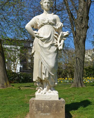 A white stone statue of a female figure in classical attire, standing on a rectangular pedestal in a grassy park with trees and yellow flowers in the background; no visible signage or text.