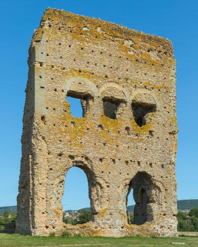 A tall, rectangular Roman brick ruin with three arched openings on the lower level and three smaller arched windows above, covered in yellow lichen, standing in a grassy field under a clear blue sky.