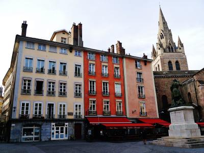 The image shows a European-style town square with colorful multi-story buildings, red awnings over ground-floor cafes, a prominent stone church tower with Gothic spires in the background, and a bronze statue on a pedestal in the foreground right.