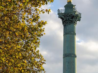 The image shows the Colonne de Juillet, a large bronze column topped with a golden winged figure of Liberty, situated in a wide public square with surrounding Haussmann-style buildings, trees, and a busy road in the foreground.