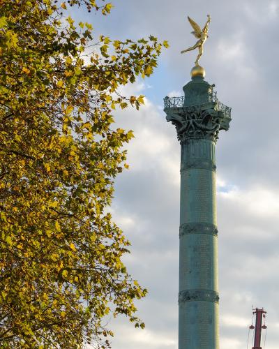 The image shows the Colonne de Juillet, a large bronze column topped with a golden winged figure of Liberty, situated in a wide public square with surrounding Haussmann-style buildings, trees, and a busy road in the foreground.
