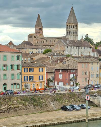 A view of the town of Tournus, France, featuring the Saint-Philibert Abbey in the background, colorful buildings along the riverbank, and a road with parked cars in the foreground.