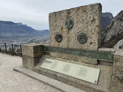 A stone monument with bronze plaques and a plaque reading "Terrasse des Géologues" overlooking a valley and mountains.
