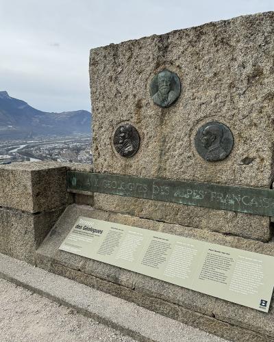 A stone monument with bronze plaques and a plaque reading "Terrasse des Géologues" overlooking a valley and mountains.