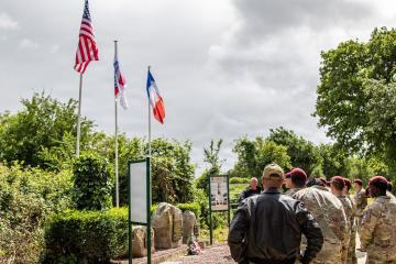 1. A group of military personnel and civilians stand outdoors near three flagpoles flying the US, French, and a third flag, with informational signs and stone markers in a green, park-like setting.