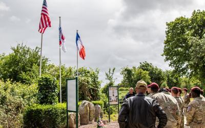 A group of military personnel and civilians stand outdoors near three flagpoles flying the US, French, and a third flag, with informational signs and stone markers in a green, park-like setting.