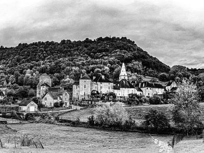 A black-and-white panoramic photograph showing a cluster of historic stone buildings, including a prominent church with a spire and fortified towers, situated at the base of a densely wooded hill in a rural valley setting.