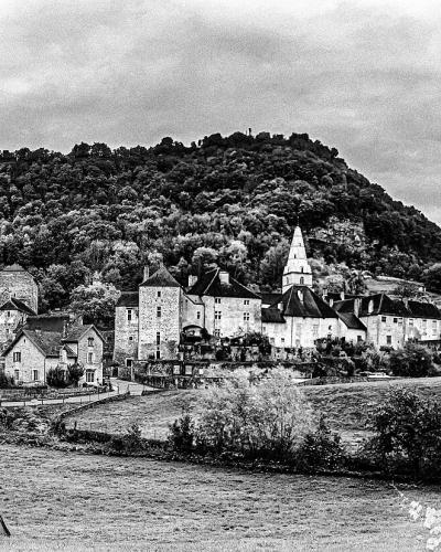 A black-and-white panoramic photograph showing a cluster of historic stone buildings, including a prominent church with a spire and fortified towers, situated at the base of a densely wooded hill in a rural valley setting.