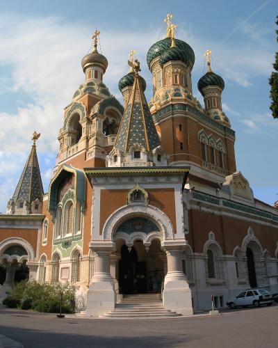 A low-angle photograph of a large Russian Orthodox cathedral with distinctive green and gold onion domes, brick and stone facade, and arched entrance, set against a blue sky with some clouds and trees on the right.