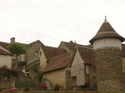 The image displays a cluster of traditional stone buildings with steep, tiled roofs, including a prominent stone tower with a conical roof on the right, set within a village environment.