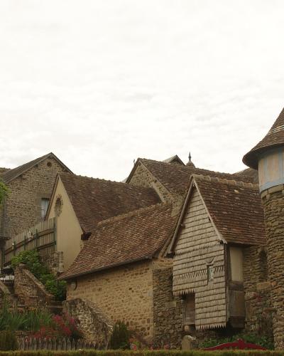 The image displays a cluster of traditional stone buildings with steep, tiled roofs, including a prominent stone tower with a conical roof on the right, set within a village environment.