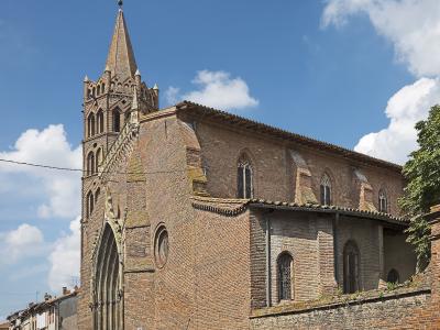 A large brick church with a tall, pointed spire and Gothic-style arched windows, set against a blue sky with white clouds, located in an urban street setting.