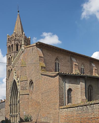 A large brick church with a tall, pointed spire and Gothic-style arched windows, set against a blue sky with white clouds, located in an urban street setting.