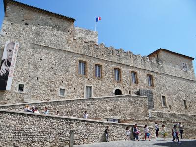 The image shows the exterior of a historic stone castle with crenellated walls, a French flag flying atop, and a large vertical banner on the left reading "Musée Picasso, Antibes"; people are walking along a paved path in front, and a small sign n