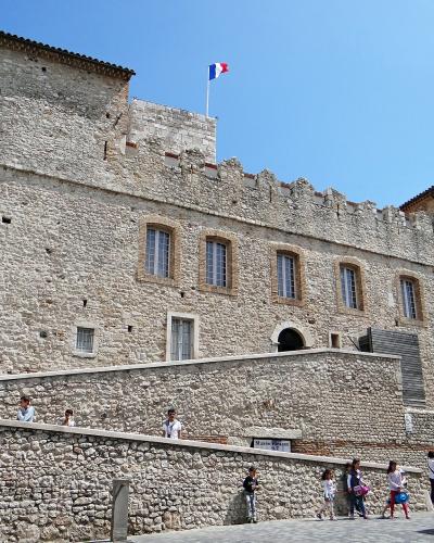 The image shows the exterior of a historic stone castle with crenellated walls, a French flag flying atop, and a large vertical banner on the left reading "Musée Picasso, Antibes"; people are walking along a paved path in front, and a small sign n
