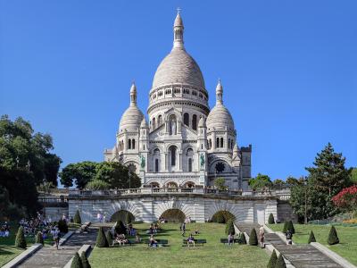 A large white basilica with a prominent central dome and two smaller flanking domes, featuring Romano-Byzantine architecture, situated atop a hill with wide staircases and terraced gardens in the foreground under a clear blue sky.
