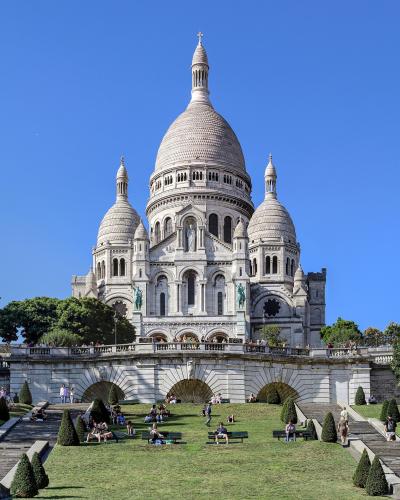 A large white basilica with a prominent central dome and two smaller flanking domes, featuring Romano-Byzantine architecture, situated atop a hill with wide staircases and terraced gardens in the foreground under a clear blue sky.