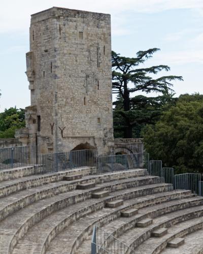 The image shows the stone seating tiers of an ancient Roman amphitheater or theater, with a tall, rectangular medieval stone tower (the Tour de l'Horloge) rising behind it, set against trees and sky.