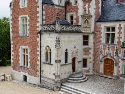 The image shows the courtyard of a historic French château featuring distinctive red brick and white stone masonry, Gothic-style arched windows, a stone balcony with tracery, and a cobblestone ground.