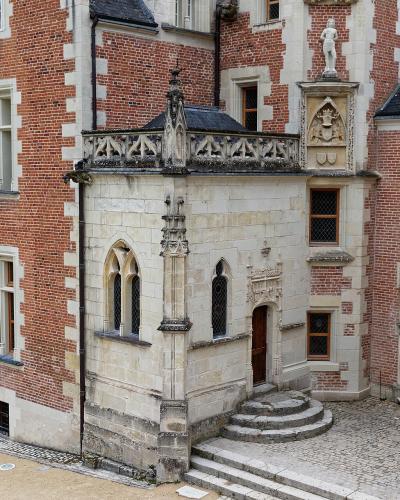 The image shows the courtyard of a historic French château featuring distinctive red brick and white stone masonry, Gothic-style arched windows, a stone balcony with tracery, and a cobblestone ground.