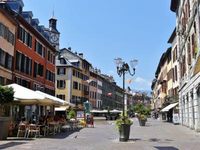 A sunlit European-style pedestrian square lined with colorful multi-story buildings featuring shutters, outdoor café seating under white umbrellas, a central ornate lamppost, and a clock tower in the background; signage includes French-language me