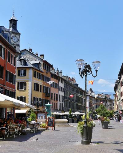 A sunlit European-style pedestrian square lined with colorful multi-story buildings featuring shutters, outdoor café seating under white umbrellas, a central ornate lamppost, and a clock tower in the background; signage includes French-language me