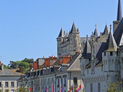 A street-level view of Saumur, France, featuring the Château de Saumur in the background, the Hôtel Cristal in the mid-ground, and pedestrians walking along a street lined with flags and parked cars.