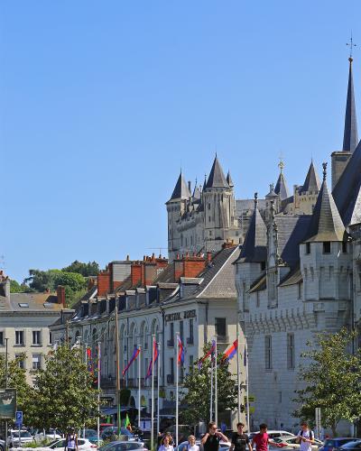A street-level view of Saumur, France, featuring the Château de Saumur in the background, the Hôtel Cristal in the mid-ground, and pedestrians walking along a street lined with flags and parked cars.