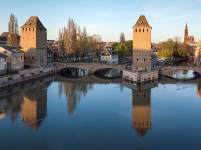 A panoramic view of the Ponts Couverts bridge complex in Strasbourg, France, featuring its three distinctive medieval towers (Tour de l'Orfèvre, Tour de la Pêche, and Tour du Faux-Rempart) spanning the Ill River, with reflections visible in the wa