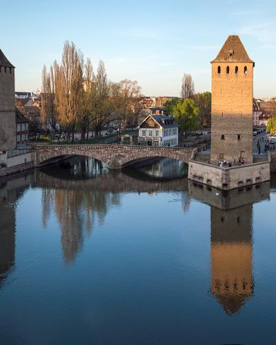 A panoramic view of the Ponts Couverts bridge complex in Strasbourg, France, featuring its three distinctive medieval towers (Tour de l'Orfèvre, Tour de la Pêche, and Tour du Faux-Rempart) spanning the Ill River, with reflections visible in the wa