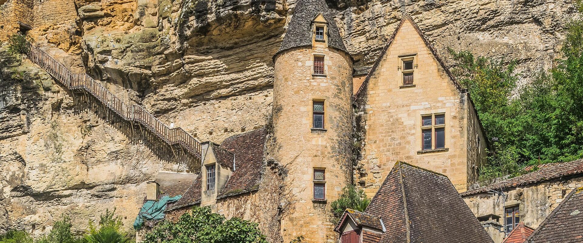 A stone castle with a conical-roofed tower and adjacent buildings nestled into a limestone cliff face, with wooden walkways and stairs built into the rock above; no visible signage or text.