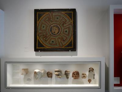 A museum interior display featuring a large square Roman mosaic with a central Medusa head and geometric border, mounted above a glass vitrine containing various Roman stone and terracotta sculptural fragments including heads and busts.