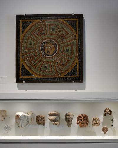 A museum interior display featuring a large square Roman mosaic with a central Medusa head and geometric border, mounted above a glass vitrine containing various Roman stone and terracotta sculptural fragments including heads and busts.
