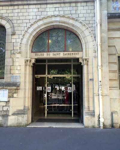 The image shows the entrance to a stone building with an arched doorway and a stained-glass window above it; the inscription "EGLISE DU SAINT SACREMENT" is carved into the lintel, and a small plaque on the left wall reads "EGLISE CATHOLIQUE"; the