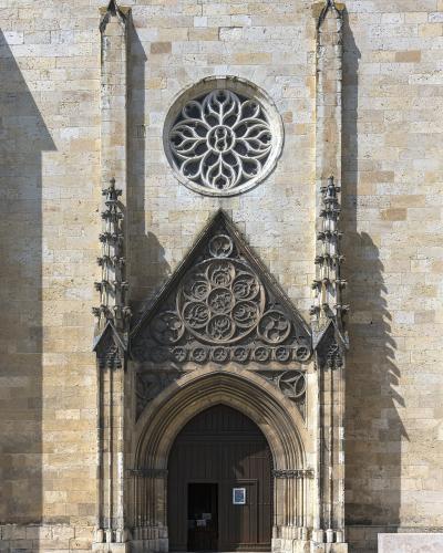 The image shows the ornate Gothic stone entrance of a cathedral, featuring a large rose window above a pointed arch doorway with intricate tracery and flanking pinnacles.