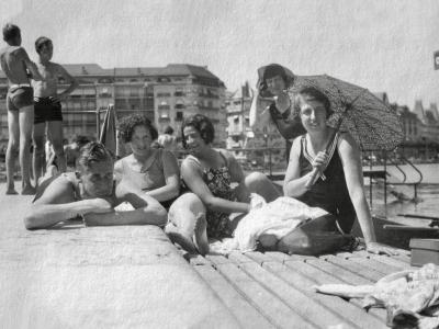 A black and white photograph from the 1930s showing a group of people in vintage swimwear relaxing on a wooden pier or jetty, with Belle Époque architecture in the background.