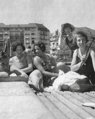 A black and white photograph from the 1930s showing a group of people in vintage swimwear relaxing on a wooden pier or jetty, with Belle Époque architecture in the background.
