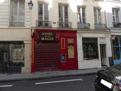 The image shows the exterior of a building with a red facade and a black sign reading "MUSEE DE LA MAGIE" above a closed metal shutter, located on a city street next to other shops.