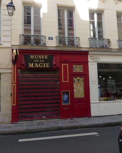 The image shows the exterior of a building with a red facade and a black sign reading "MUSEE DE LA MAGIE" above a closed metal shutter, located on a city street next to other shops.