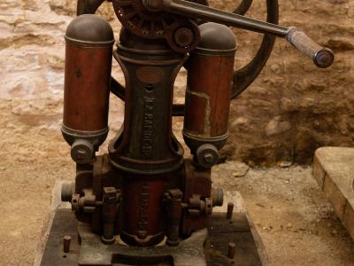 An antique red and black cast-iron wine pump with a large handwheel, displayed on wooden blocks against a rough stone wall, featuring a bilingual label reading "POMPE A SOUTIRER LE VIN / PUMP TO EXTRACT THE WINE".