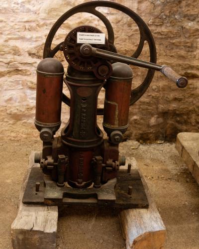 An antique red and black cast-iron wine pump with a large handwheel, displayed on wooden blocks against a rough stone wall, featuring a bilingual label reading "POMPE A SOUTIRER LE VIN / PUMP TO EXTRACT THE WINE".