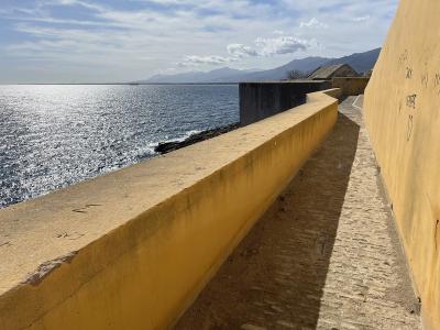 A view from a paved walkway along a high yellow wall overlooking the sea, with mountains in the distance.