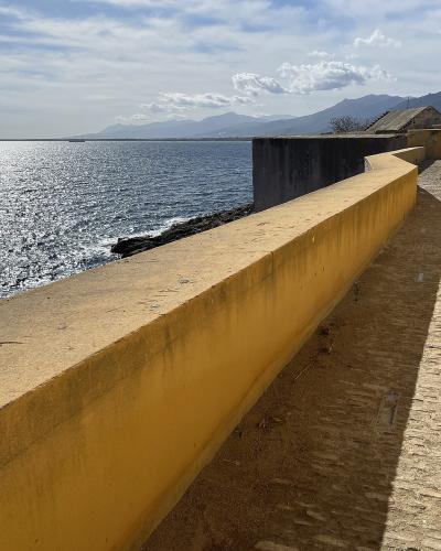 A view from a paved walkway along a high yellow wall overlooking the sea, with mountains in the distance.