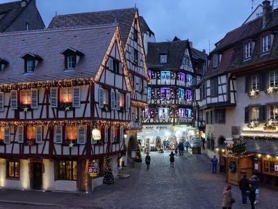 The image shows a cobblestone street in a historic European town at dusk, featuring half-timbered buildings decorated with Christmas lights and festive displays, including a shop sign reading "VIEUX PINON" and a sign for "DE GERMANY".