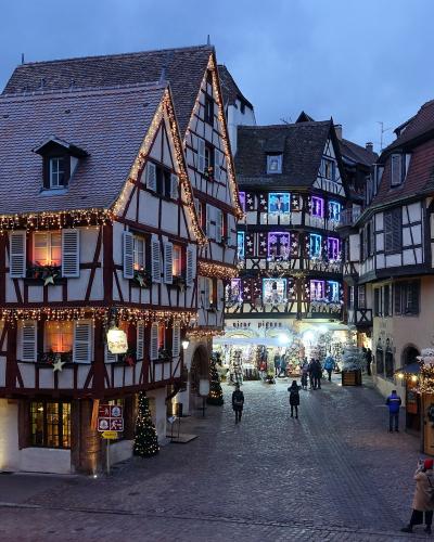 The image shows a cobblestone street in a historic European town at dusk, featuring half-timbered buildings decorated with Christmas lights and festive displays, including a shop sign reading "VIEUX PINON" and a sign for "DE GERMANY".