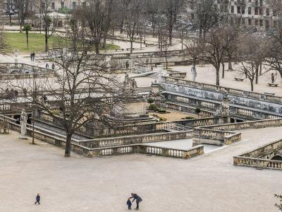 A high-angle view of a large, terraced historic park featuring extensive stone balustrades, multiple levels of fountains and basins, classical statues, and bare deciduous trees with people walking on gravel paths.