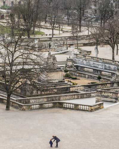 A high-angle view of a large, terraced historic park featuring extensive stone balustrades, multiple levels of fountains and basins, classical statues, and bare deciduous trees with people walking on gravel paths.