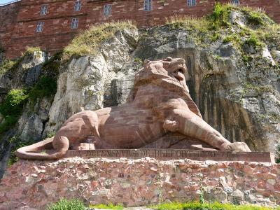 A close-up profile view of a massive sandstone lion sculpture carved directly into a rock face, showing the head, mane, and part of the neck against a plain sky.