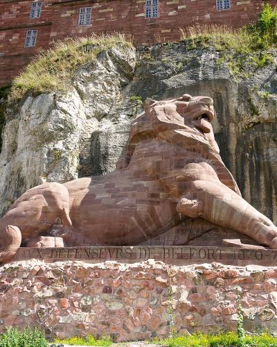 A close-up profile view of a massive sandstone lion sculpture carved directly into a rock face, showing the head, mane, and part of the neck against a plain sky.