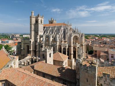 A high-angle photograph of a large, unfinished Gothic cathedral with a prominent square bell tower on the left, flying buttresses, and a terracotta-tiled roof, set within a dense urban environment of similar tiled rooftops under a blue sky.